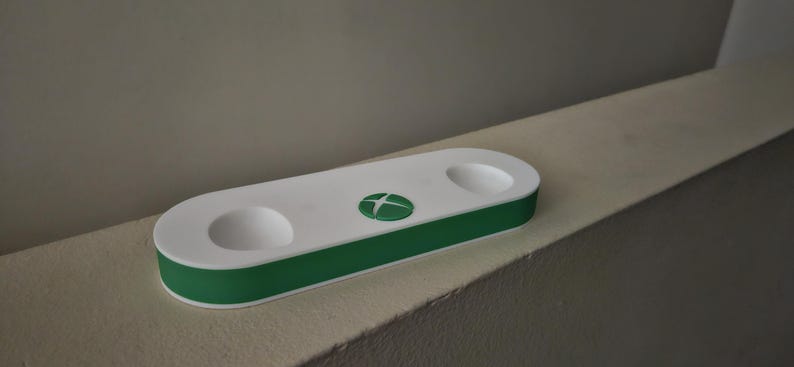 K&ouml;nnte beinhalten: Eine wei&szlig;-gr&uuml;ne Controller-Ladestation in Pillenform mit zwei runden, vertieften Aussparungen f&uuml;r Controller und einem mittigen gr&uuml;nen runden Logo mit einem wei&szlig;en, vierquadrantigen Symbol auf der Oberseite.