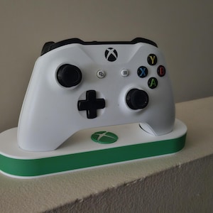 May include: A white Xbox wireless controller resting on a white and green charging dock. The controller features black analog sticks, a black directional pad, and the standard colorful A, B, X, and Y buttons. The Xbox logo is visible on the controller.