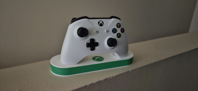K&ouml;nnte beinhalten: Ein wei&szlig;er kabelloser Xbox-Controller auf einer wei&szlig;en und gr&uuml;nen Ladestation. Der Controller verf&uuml;gt &uuml;ber schwarze Analogsticks, ein schwarzes Steuerkreuz sowie die farbigen A-, B-, X- und Y-Tasten. Das Xbox-Logo ist auf dem Controller zu sehen.