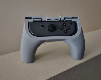 Könnte beinhalten: Ein grauer Kunststoff-Controller-Griff, der eine dunkelgraue Controller-Komponente umschließt. Der Griff verfügt über ein geschwungenes, ergonomisches Design mit einem zentralen Schlitz für den Controller, der einen schwarzen Joystick und vier runde Aktionstasten enthält.