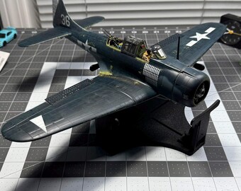 Könnte beinhalten: Ein dunkelblaues Modellflugzeug eines Sturzkampfbombers aus dem Zweiten Weltkrieg mit der Nummer 36 auf dem Leitwerk und Rumpf, weißen Sternabzeichen auf den Flügeln, einem detaillierten Cockpit mit Pilotfigur und einem schwarzen Ständer.