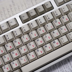 May include: A white keyboard with black keys and red symbols. The keys have various symbols and letters, including the alphabet, numbers, and punctuation marks. The keyboard has a silver frame and is sitting on a piece of paper.