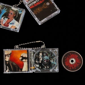 May include: Clear plastic CD cases with various album art, some attached to keychains. One case shows a woman in a nurse's uniform. Another displays the text "SYSTEM OF A DOWN MEZMERIZE". A loose CD with a red and black design is also present.