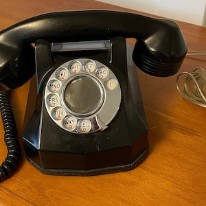 Puede incluir: Un teléfono de disco negro con una cara plateada y un cable negro en espiral. El teléfono tiene un dial plateado con números y letras negras. El teléfono está sobre una superficie de madera.