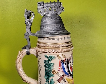 May include: A ceramic beer stein with a pewter lid featuring a train. The stein is decorated with a green wreath, flags, and a train. The text "Bundesbahn Abteilung der Verkehrs" is visible on the stein.