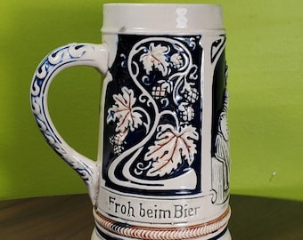 Op de afbeelding: Een witte keramische bierkan met blauwe en bruine accenten. De kan heeft een wijnstok met bladeren en druiven, en de tekst "Froh beim Bier".