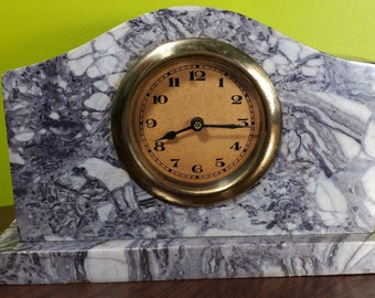 Könnte beinhalten: Eine Marmor-Kaminuhr mit goldfarbenem Rahmen und beigem Zifferblatt. Der Marmor der Uhr hat ein grau-weißes Muster. Die Uhr steht auf einer Holzoberfläche.