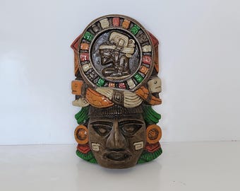 May include: A colorful, hand-painted, ceramic wall mask depicting a Mayan deity with a circular headpiece and intricate details. The mask is brown, green, orange, and white.