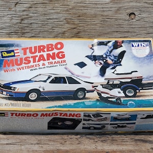 May include: A model kit box for a white and blue 1978 Ford Mustang Turbo with a trailer and two jet skis. The box features the text "Turbo Mustang" and "With Wetbikes & Trailer".