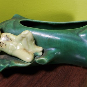 以下が含まれることがあります： 葉の形をした緑色のセラミック製のプランターに、小さく緑と黄色の妖精の置物が横たわっています。妖精は、目を閉じ、笑顔のペイントされた顔をしています。