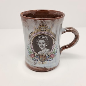 May include: A white ceramic mug with a brown rim and handle. The mug features a printed image of Queen Elizabeth II with the text "Queen Elizabeth II Silver Jubilee 1952 - 1977".