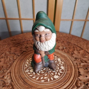 Könnte beinhalten: Eine grüne Keramik-Gartenzwergfigur mit weißem Bart und roten Hosen, die eine Schaufel hält. Der Zwerg steht auf einem runden Holzsockel mit einem geschnitzten Blumendesign.