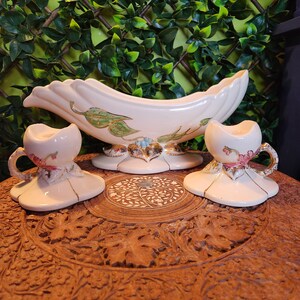 Puede incluir: Un juego de tres candelabros de cerámica vintage de color crema con detalles dorados. La pieza central es un cuenco grande y curvo con un diseño floral. Las dos piezas más pequeñas son candelabros con un diseño floral y una pequeña asa.