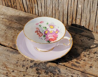 Puede incluir: Un juego de taza y platillo de té de color lavanda con un diseño floral en tonos rosa, morado y amarillo. La taza y el platillo tienen un borde dorado.