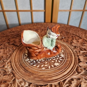 Könnte beinhalten: Eine Keramikfigur eines Elfen, der in einem braunen Schuh sitzt. Der Elf trägt ein grünes Hemd und eine rote Mütze. Der Schuh ist mit weißen und braunen Details verziert.