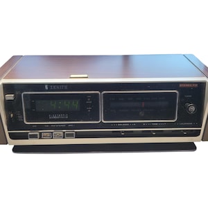 Puede incluir: Radio reloj Zenith vintage con tapa y panel frontal de imitación madera. El reloj digital muestra las 4:44. La radio tiene un sintonizador FM estéreo y un control de balance. El radio reloj tiene una base negra y detalles dorados.