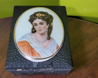 Puede incluir: Una caja de madera negra con una tapa de porcelana ovalada blanca que presenta un retrato pintado de una mujer con cabello oscuro y un collar de perlas. La mujer lleva un vestido blanco y naranja.
