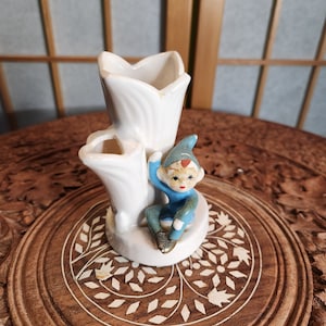 Könnte beinhalten: Eine weiße Keramikvase mit drei Öffnungen, geformt wie Tulpenblätter. Eine kleine, skurrile Figur eines blau gekleideten Elfen mit spitzer Mütze sitzt auf dem Sockel. Die Vase steht auf einer geschnitzten Holzoberfläche.