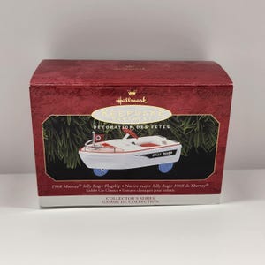 May include: A red and white Hallmark Keepsake ornament featuring a 1968 Murray Jolly Roger Flagship Kiddie Car Classic. The ornament is in its original box, which features the Hallmark logo and the text "Keepsake Ornament" and "Decoration des Fetes".