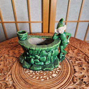 Könnte beinhalten: Ein grüner Keramik-Brunnen-Pflanztopf mit einem kleinen Eimer oben und einer grünen Elfenfigur, die sich an die Seite lehnt.