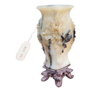 Könnte beinhalten: Eine geschnitzte, cremefarbene Vase mit floralen und Vogeldetails, montiert auf einem dekorativen, dunkelrotbraunen Ständer. Die Vase hat ein Etikett mit der Nummer "208-27". Die Vase ist wahrscheinlich aus Stein oder einem ähnlichen Material.