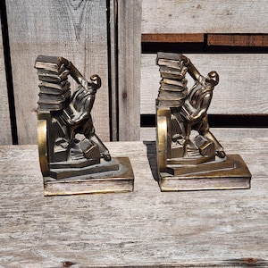 Könnte beinhalten: Ein Paar antike Bronze-Buchstützen mit einer Figur, die einen Stapel Bücher hält. Die Buchstützen sind in gutem Zustand mit einigen Gebrauchsspuren.