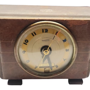 Op de afbeelding: Vintage bruine houten klok met een crèmekleurige wijzerplaat en gouden accenten. De wijzerplaat toont de tijd en de dag. De klok is gemarkeerd met "HAMMOND" en heeft een rechthoekige vorm. Ongeveer 15 cm breed.
