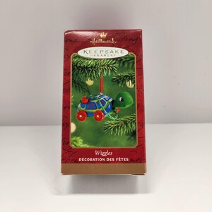 Puede incluir: Una caja roja con un adorno Hallmark Keepsake en su interior. El adorno es una tortuga azul y verde con ruedas rojas. La caja dice "Wiggles Décoration des Fêtes".