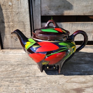 Puede incluir: Una tetera marrón con un diseño floral rojo, amarillo y verde. La tetera tiene una tapa y un pico. La tetera está sentada sobre una superficie de madera.