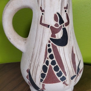 May include: A white ceramic pitcher with a brown, black, and white stylized design of a person dancing. The pitcher has a handle and a rounded body.