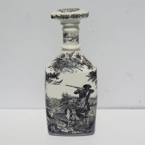 Op de afbeelding: Een witte keramische karaf met een zwart-wit jachtscène-ontwerp. De karaf heeft een stop en een gedetailleerde illustratie van een jager, honden en vogels in een landschap.