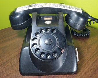 May include: A black rotary dial phone with a black coiled cord. The phone has a black dial with white numbers 1 through 9 and 0. The phone has a black handset with a black cord.