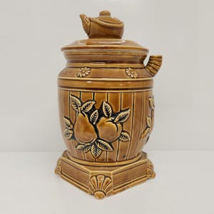 Puede incluir: Una tetera de cerámica marrón con tapa y boquilla. La tetera tiene un diseño en forma de barril con líneas verticales y patrones de frutas y flores en relieve. La base tiene una forma octogonal decorativa.