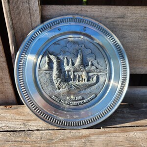 May include: A silver circular dish with a raised design of a boat and buildings. The dish has a silver rim with a raised edge. The text "THAILAND BEST HAND CRAFTED PEWTER" is engraved on the dish.