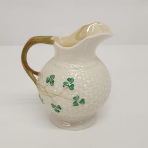 Puede incluir: Jarra pequeña de cerámica blanquecina con textura tejida. El asa es marrón y verde. Diseños de tréboles verdes pintados en la parte delantera. La jarra tiene un cuerpo redondeado y un pico acampanado.