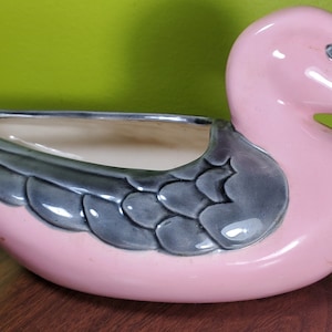 Puede incluir: Un macetero de cerámica rosa en forma de pato con un diseño de ala glaseada gris. El macetero tiene una abertura ancha para plantar.