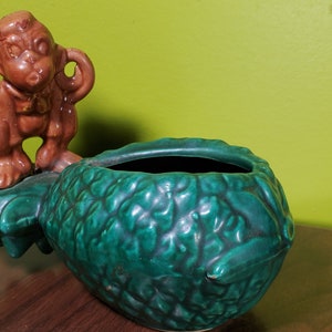 May include: A green ceramic pineapple planter with a brown monkey figurine on top. The planter has a textured surface that resembles pineapple skin.