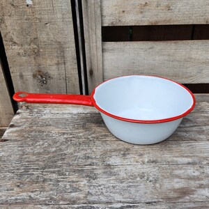 Könnte beinhalten: Ein weißer Emaille-Topf mit rotem Rand und rotem Griff. Der Topf steht auf einer Holzoberfläche.