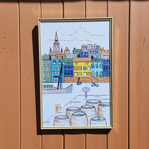 Op de afbeelding: Geborduurd kunstwerk dat een kleurrijke waterkantscène toont met gebouwen in blauw, geel, groen en wit. Een zeilboot en verschillende vaten staan op de voorgrond. Het kunstwerk is ingelijst in een goudkleurige lijst.