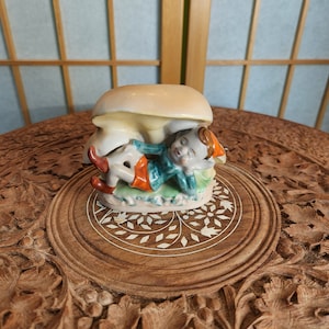 Könnte beinhalten: Eine Keramikfigur eines schlafenden Elfen, der auf einem Pilz liegt. Der Elf trägt ein grünes und orangefarbenes Outfit und einen roten Hut. Der Pilz ist weiß mit braunen Flecken.