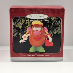 Può includere: Un ornamento Hallmark Keepsake con Mrs. Potato Head. L'ornamento è a forma di patata marrone con cappello giallo, orecchini rosa, scarpe verdi e borsetta rossa. La scatola è rossa con testo dorato.