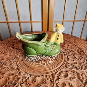 Könnte beinhalten: Ein grüner Keramik-Stiefel-Pflanztopf mit einer gelben Elfenfigur darauf. Der Elf hat schwarze Augen und einen schwarzen Mund. Der Stiefel hat eine weiße Inschrift mit der Aufschrift "c.u."