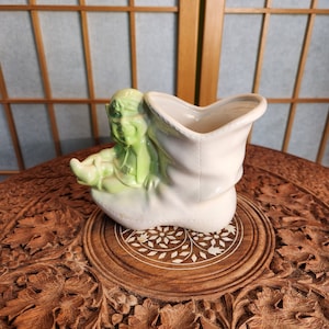 Könnte beinhalten: Eine grüne Keramik-Gnomfigur, die in einem weißen Keramik-Stiefel-Pflanztopf sitzt. Der Stiefel hat eine breite Öffnung oben und ist mit einem weißen Rand verziert.