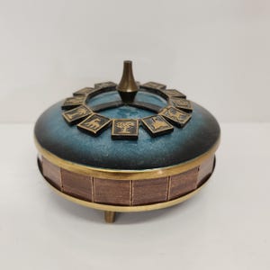 May include: A decorative box with a teal blue lid and a wooden base. The lid is adorned with bronze-coloured square tiles featuring animal and plant illustrations. A bronze-coloured finial sits atop the lid, and the base has bronze trim.