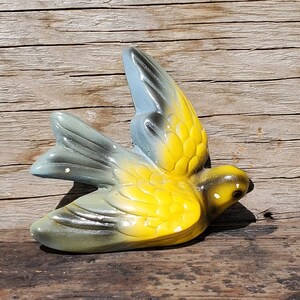 May include: A yellow and gray ceramic bird figurine with a black eye. The bird is in flight and has its wings spread out.