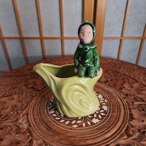Könnte beinhalten: Vintage Keramik-Pflanzgefäß in Form eines Baumstamms, hellgrün, mit einer kleinen Figur eines grün gekleideten Elfen. Der Elf hat ein rosa Gesicht und grüne Augen. Der Pflanztopf steht auf einem geschnitzten Holztisch.