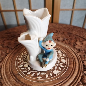 Könnte beinhalten: Eine weiße Keramikvase mit drei Öffnungen in Form einer Blume, mit einer blauen und weißen Elfenfigur, die auf der Basis sitzt.