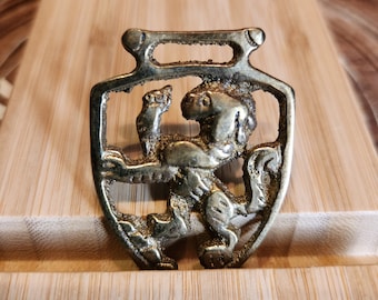 Puede incluir: Un colgante de latón con un león en un marco en forma de escudo. El león está representado en una pose estilizada y heráldica.