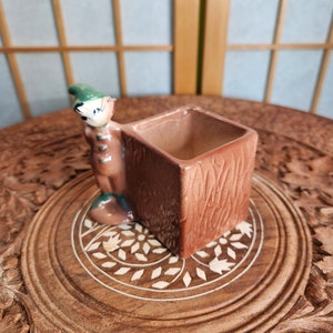 Könnte beinhalten: Ein kleiner Keramik-Pflanztopf in Form eines Baumstamms mit einer skurrilen Comicfigur, die hinter ihm hervorlugt. Der Topf ist braun mit Holzmaserung und hat oben eine quadratische Öffnung zum Pflanzen.