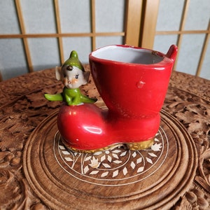 Könnte beinhalten: Ein roter Keramik-Stiefel-Pflanztopf mit einer grünen Elfenfigur darauf. Der Stiefel hat einen goldenen Rand und der Elf hat ein weißes Gesicht mit grünen Haaren und einem grünen Outfit.